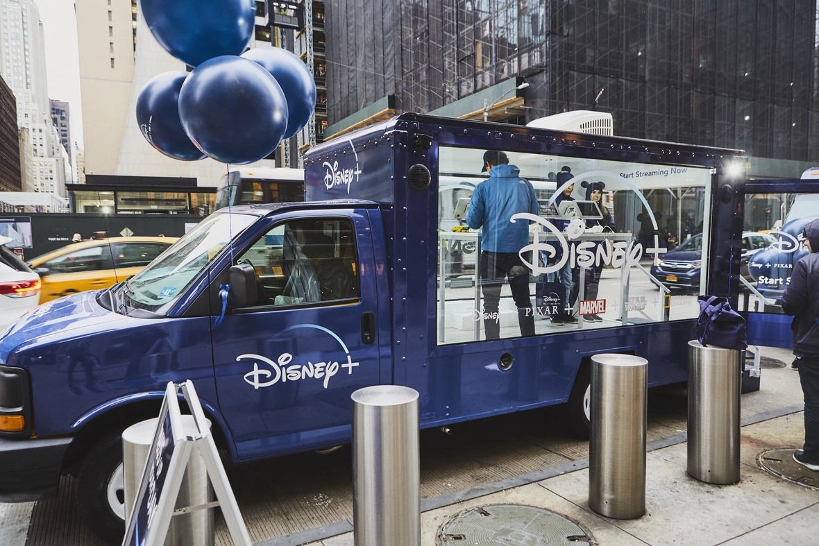 DisnDisney Mobile Showroom for collecting survey dataey Mobile Showroom for collecting survey data Disney+ branded mobile showroom featuring staff collecting survey data, surrounded by blue balloons, set in an urban environment, illustrating guerrilla marketing strategies.