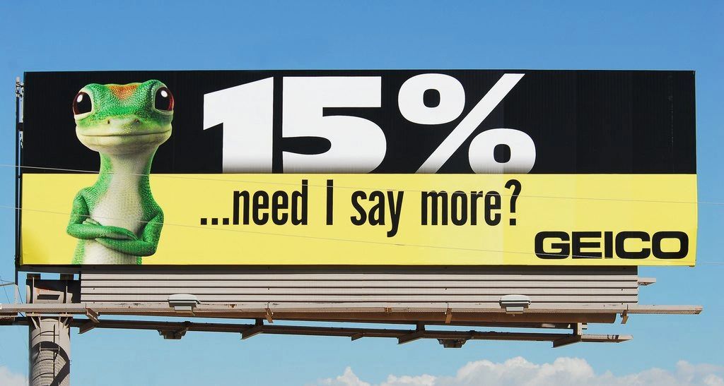 Billboard featuring a green gecko character promoting a 15% discount with the text "...need I say more?" and the GEICO logo, illustrating modern insurance advertising strategies.