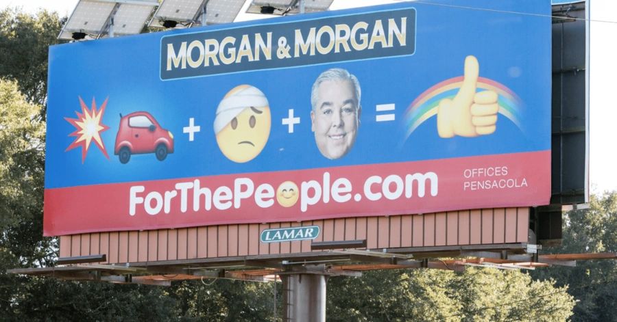 Billboard advertisement for Morgan & Morgan featuring visual symbols of a car crash, an injured person, and a smiling attorney, promoting legal services with the slogan "ForThePeople.com" in a high-traffic area.