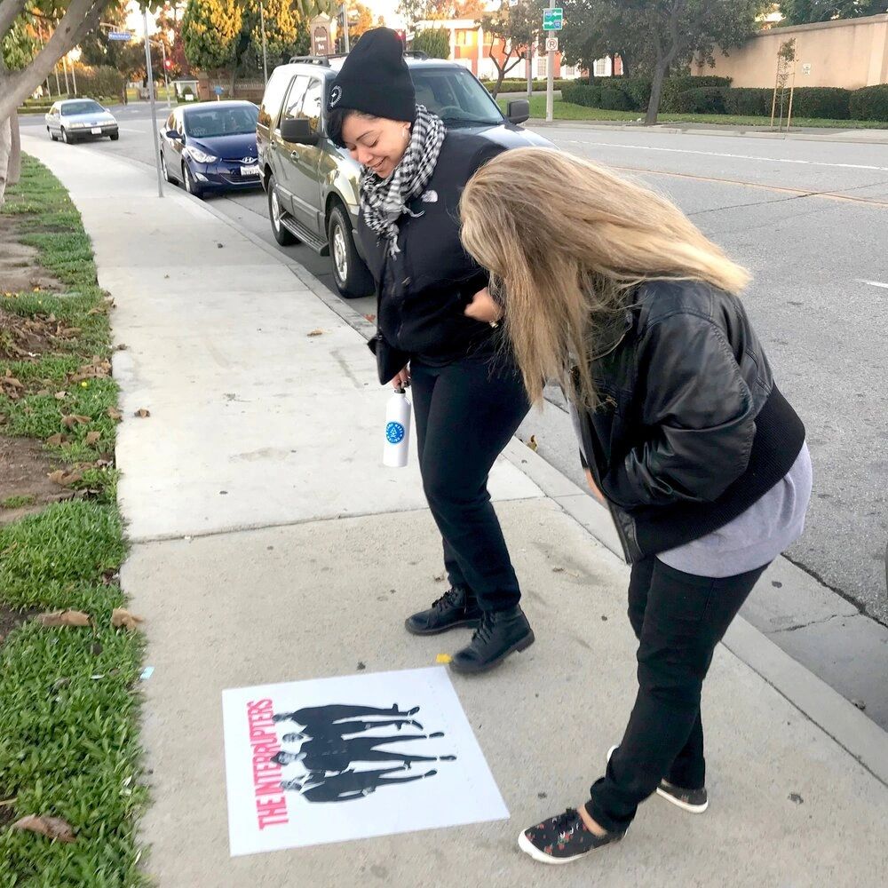 A successful sidewalk stencil marketing campaign begins with meticulous research, taking into account your brand's values, target audience perception, and local regulations. Two women interacting with a sidewalk stencil featuring a black-and-white illustration of a band, "The Interrupters," highlighting community engagement in sidewalk stencil marketing.