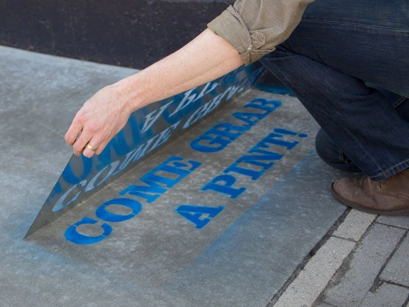 Person applying a stencil to a sidewalk, revealing the text "COME GRAB A PINT!" in blue, illustrating creative guerrilla marketing tactics.