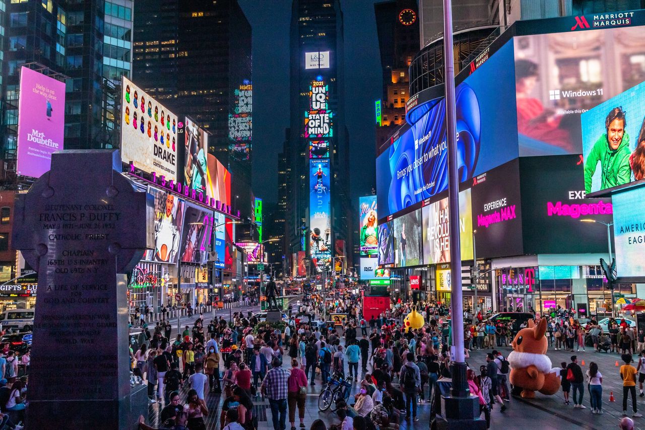Crowd of pedestrians in Times Square surrounded by vibrant digital billboards featuring advertisements for Drake, Windows 11, and augmented reality experiences, with a statue of Francis P. Duffy in the foreground.