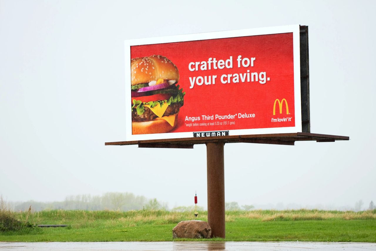 The choice between highway and urban billboard locations ultimately depends on the campaign's goals, target audience, and message strategy. Billboard advertising featuring McDonald's Angus Third Pounder Deluxe burger, with the tagline "crafted for your craving" and "I'm lovin' it," set in an outdoor environment.