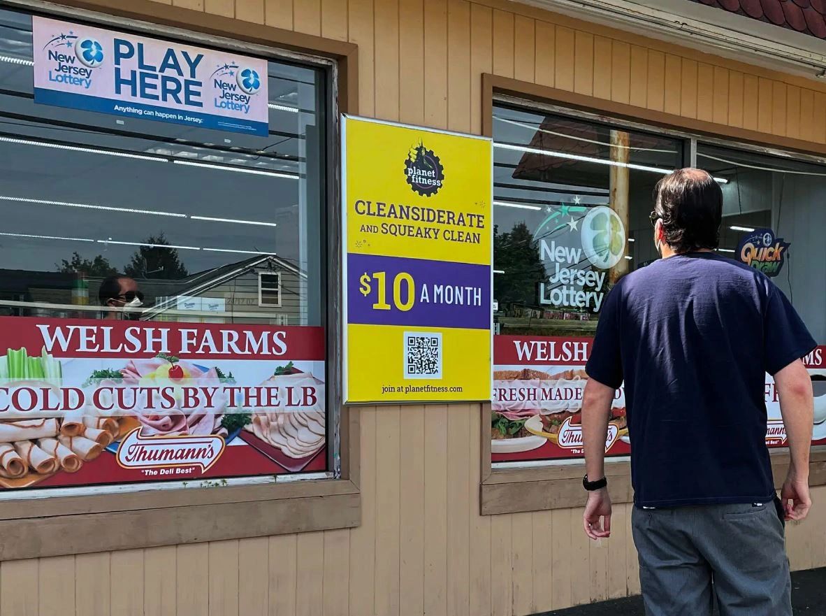 Man standing in front of convenience store window displaying advertisements for New Jersey Lottery, Welsh Farms cold cuts, and Planet Fitness membership promotion for $10 a month.