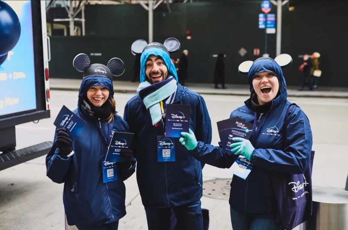 Disney brand ambassadors smiling and holding promotional materials for Disney+, wearing matching blue jackets and Mickey Mouse ear hats, in a vibrant urban setting.