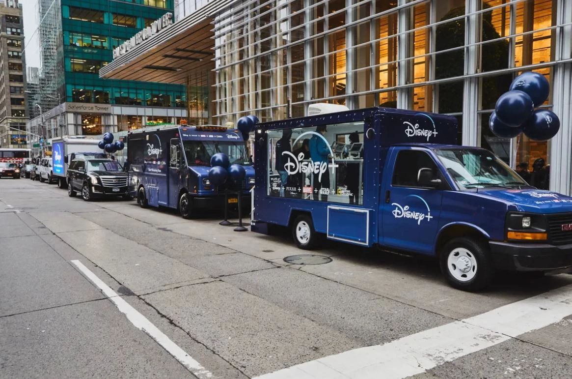 Disney+ branded food trucks lined up on city street, featuring blue color scheme, promotional signage, and balloons, showcasing guerrilla marketing activation for brand engagement.
