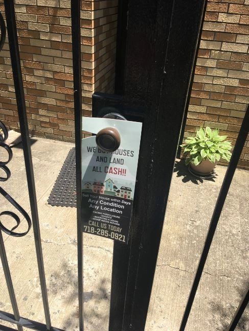 Door hanger on a gate promoting cash offers for houses and land, featuring a bold headline and contact information for potential sellers.