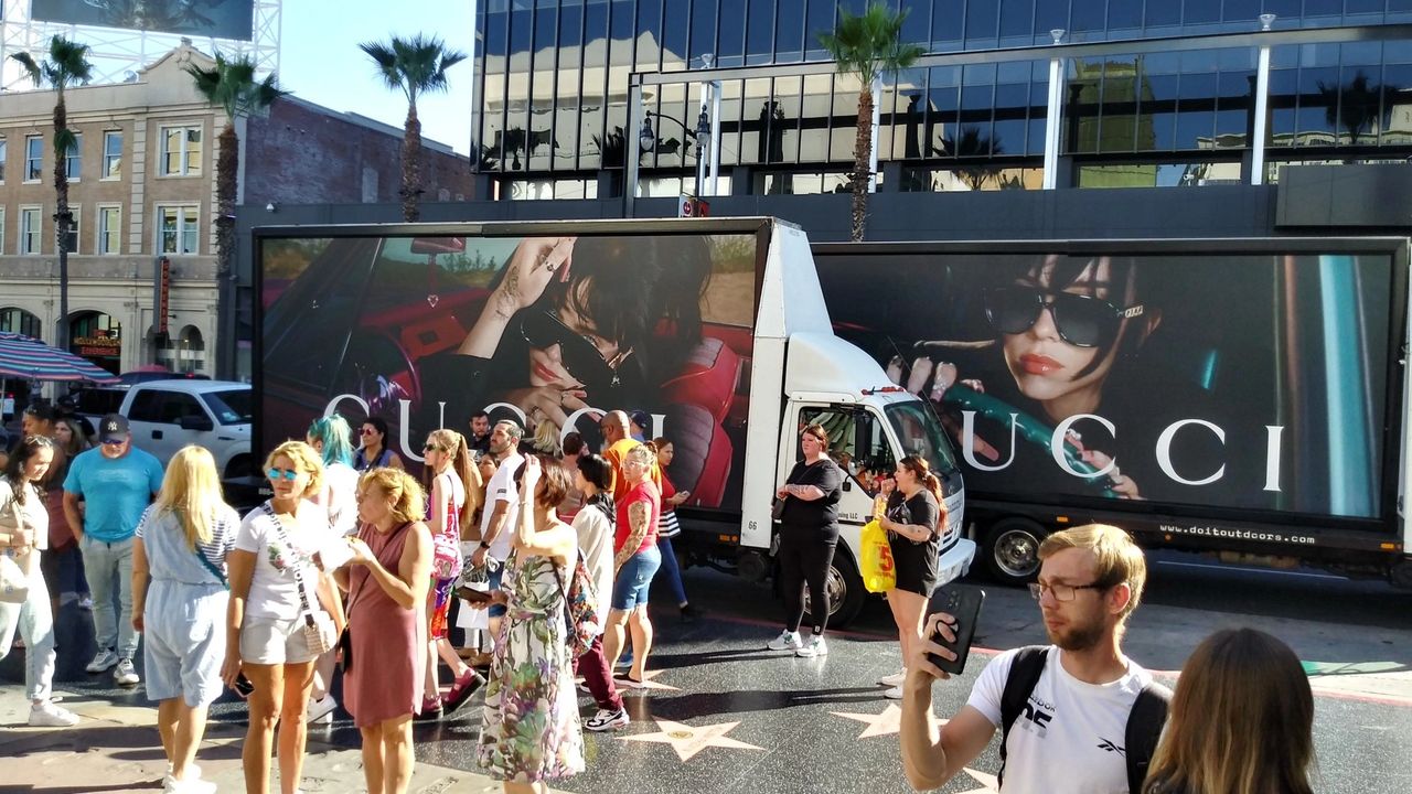 Gucci advertisement on truck featuring a model in sunglasses, surrounded by a crowd in a busy urban setting, emphasizing fashion marketing and brand identity.