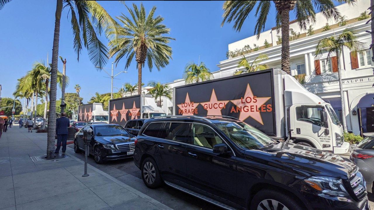 Billboard trucks displaying "LOVE GUCCI PARADE LOS ANGELES" advertisement amidst palm trees and parked vehicles in a busy urban setting, illustrating mobile advertising strategies.