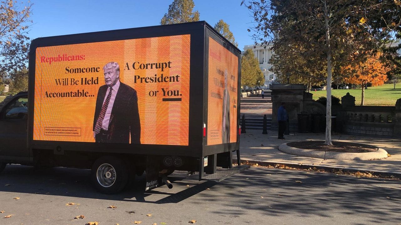 Truck displaying political advertisement featuring text about accountability and corruption, with an image of a man in a suit, set against an autumn backdrop.