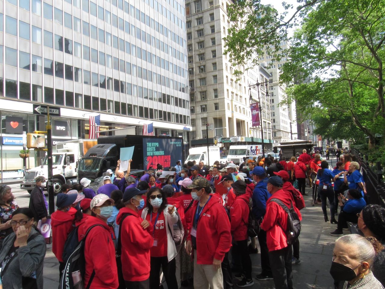Crowd gathered for protest in New York City, featuring LED billboard truck displaying social justice message, participants in red and blue attire, and urban backdrop.