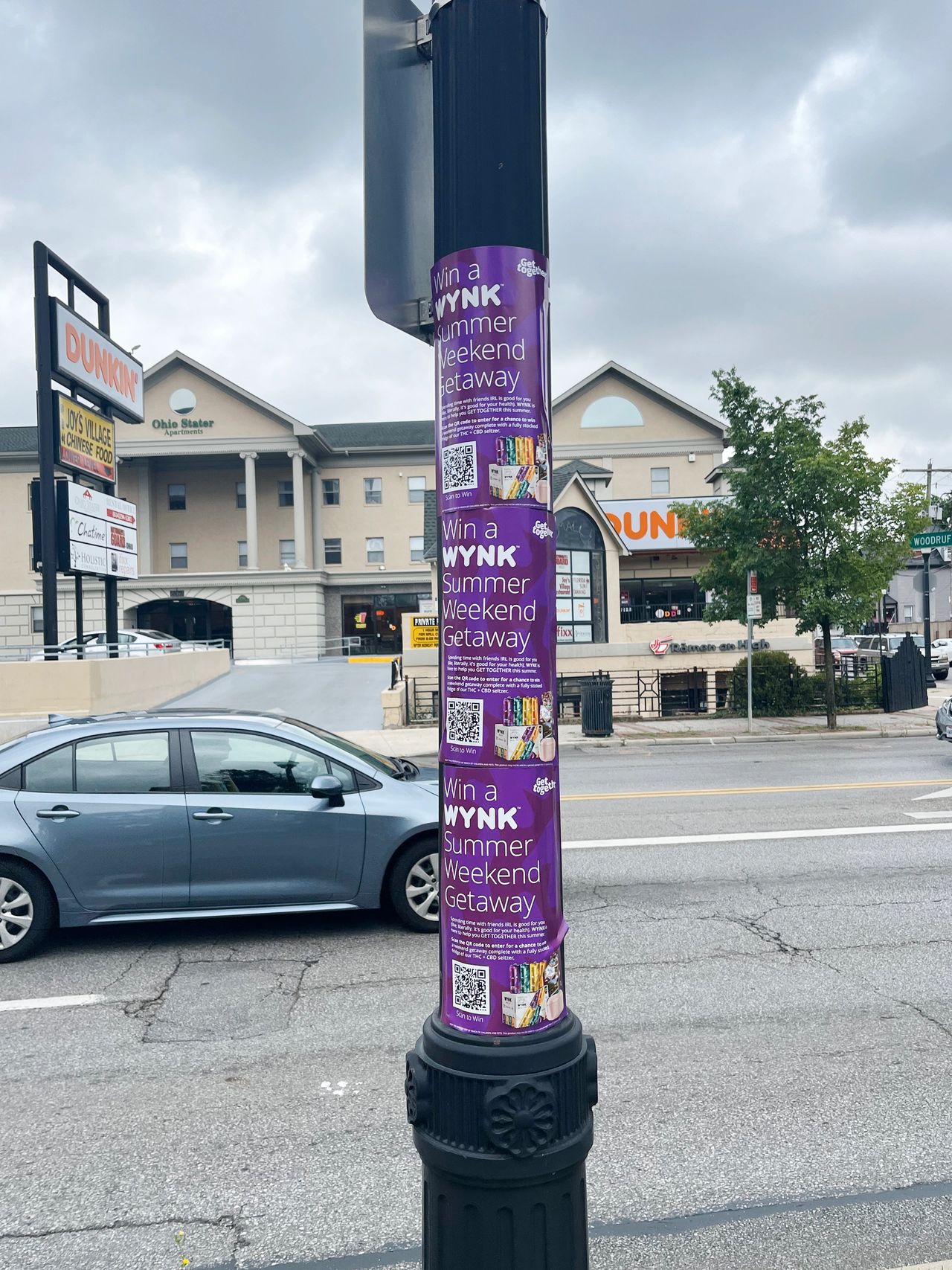 Purple promotional poster on a streetlight advertising a "WYNK Summer Weekend Getaway," featuring QR codes and colorful graphics, with a Dunkin' store and apartment building in the background, highlighting urban marketing strategies.