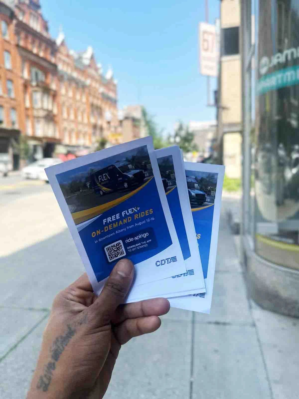 Hand holding flyers promoting "Free FLEX On-Demand Rides" service in Burlington, Vermont, with a city street and historic buildings in the background, emphasizing brand activation and community engagement.