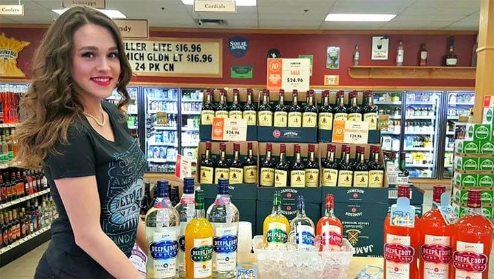 Woman promoting liquor products at a sampling event, showcasing colorful bottles of Deep Eddy vodka and various mixers, with a display of whiskey in the background, emphasizing brand experience and customer engagement.