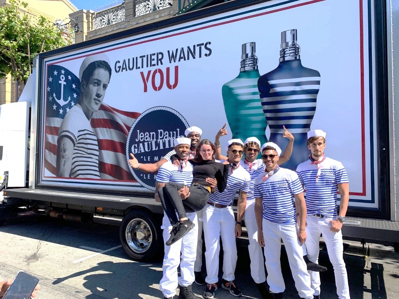 Mobile billboard promoting Jean Paul Gaultier's fragrance, featuring a vintage design with models in striped attire, set against an American flag backdrop, surrounded by a group of enthusiastic promoters.