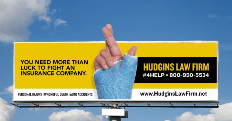 Billboard advertisement for Hudgins Law Firm featuring a hand with a bandaged finger crossing for luck, emphasizing the message "You need more than luck to fight an insurance company," highlighting personal injury legal services.