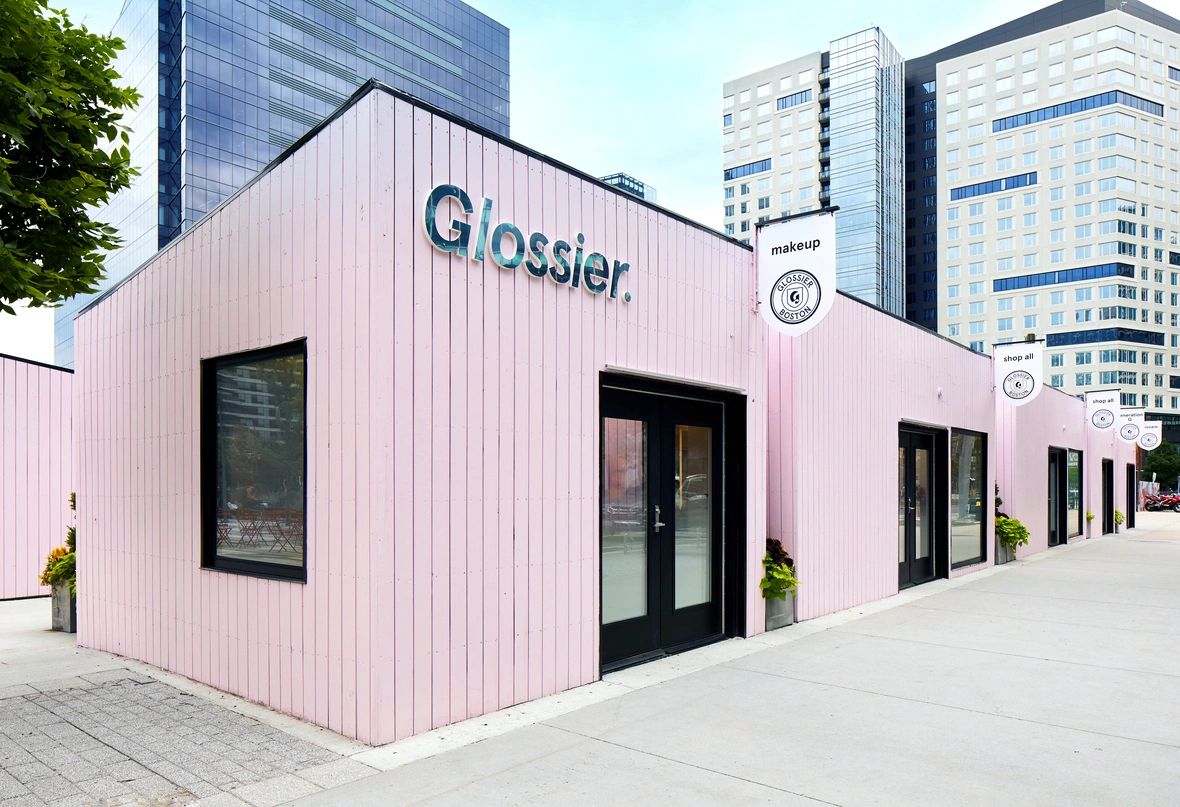 Glossier pop-up store with pink wooden exterior and black-framed windows, located in an urban setting, showcasing signage for makeup and shop options.