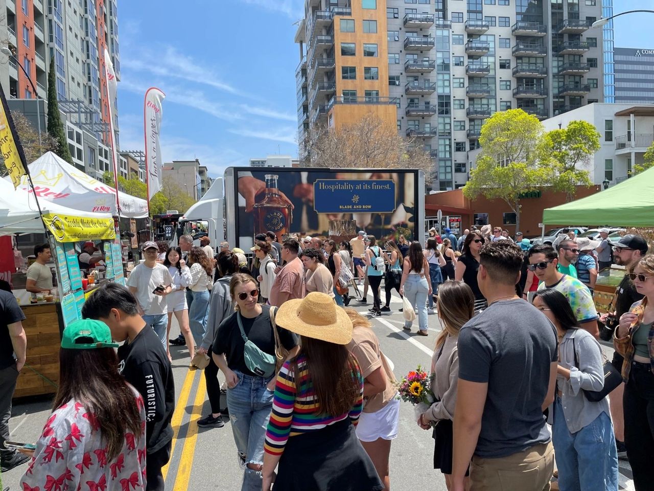 Marketing activations are strategic campaigns and initiatives designed to engage consumers and generate excitement about a brand or product. Crowd enjoying a vibrant outdoor market with food stalls and promotional billboard featuring "Hospitality at its finest" for Blade and Bow whiskey, emphasizing engaging marketing activations.