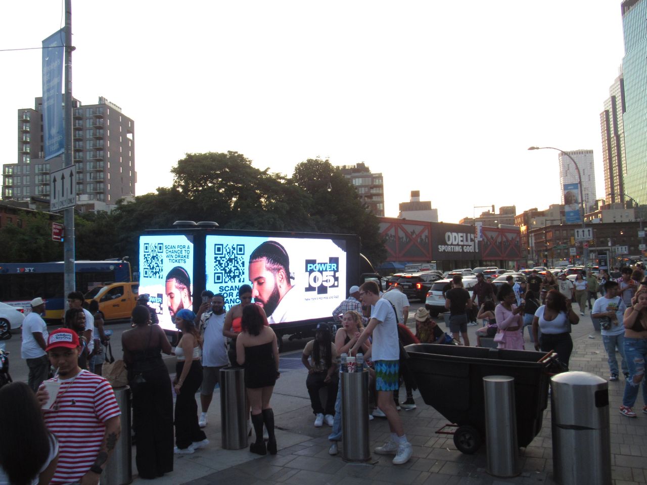 Crowd gathered around a promotional truck displaying a QR code and advertisement for a contest to win Drake tickets, with urban setting and evening light.