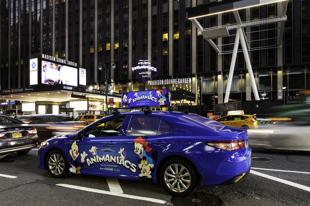 Blue taxi featuring "Animaniacs" advertisement parked near Madison Square Garden, illuminated billboards in background, showcasing urban advertising strategies.