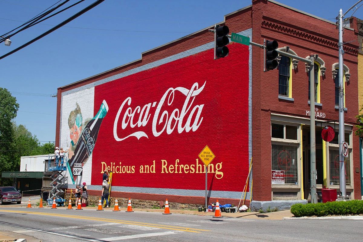 Coca-Cola mural painting featuring the iconic logo and slogan "Delicious and Refreshing," with artists working on a vibrant red wall, showcasing mural art as a powerful marketing strategy.
