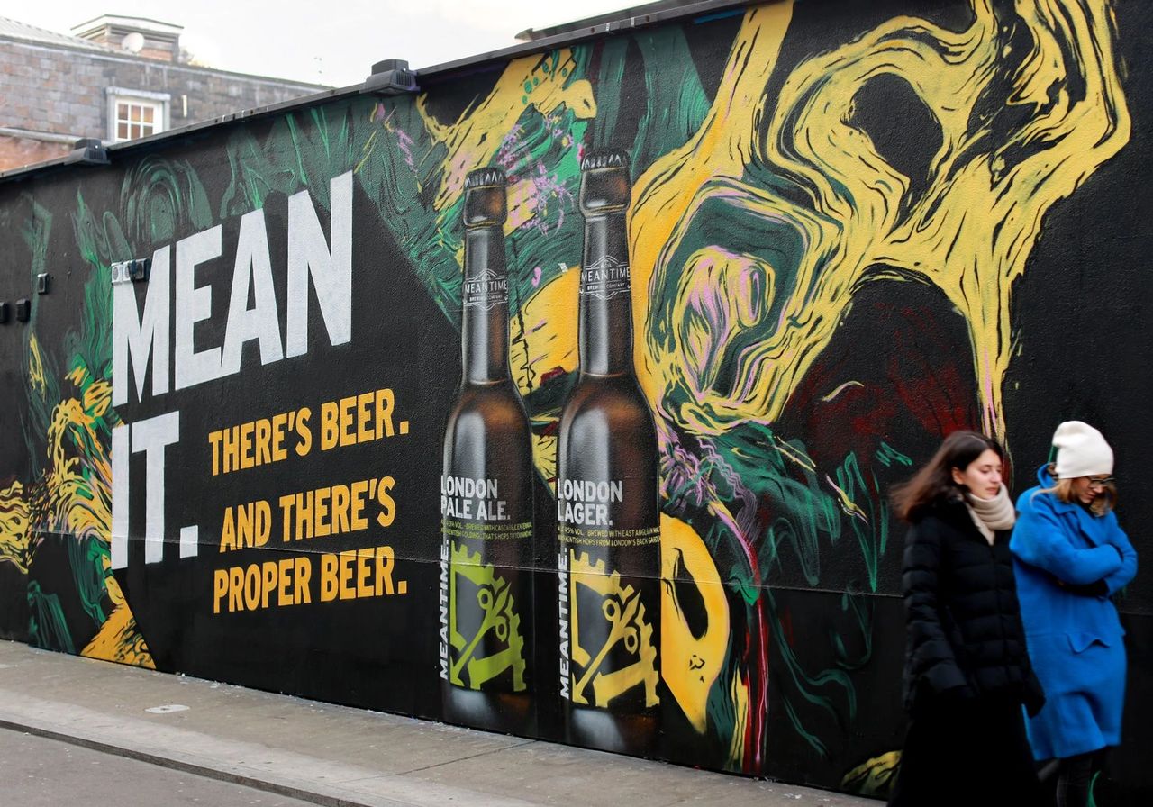 Mural painting featuring bold text "MEAN IT. THERE'S BEER. AND THERE'S PROPER BEER." alongside images of beer bottles, vibrant colors, and abstract designs, illustrating the intersection of art and guerrilla marketing in urban spaces.