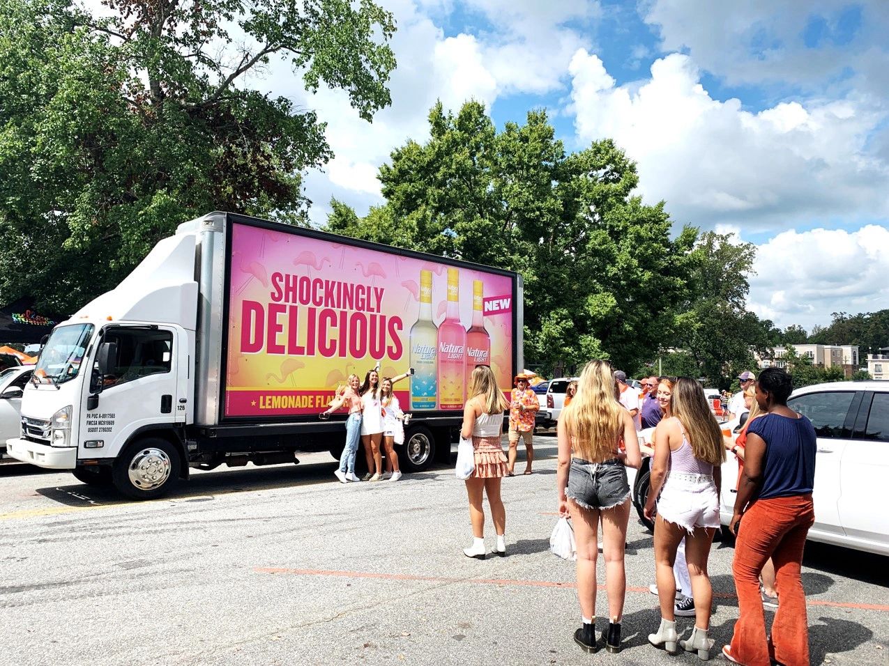 Billboard truck advertising Natty Vodka with "Shockingly Delicious" and "Lemonade Flavor" signage, surrounded by a crowd of young people engaging with the promotion.