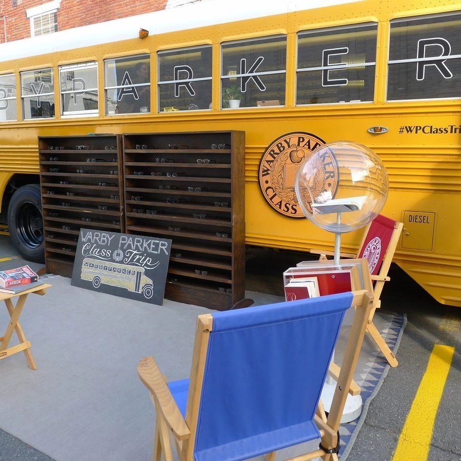 Warby Parker pop-up shop featuring a vintage yellow bus, display of eyeglasses, and inviting seating area, emphasizing immersive brand experience and consumer engagement.