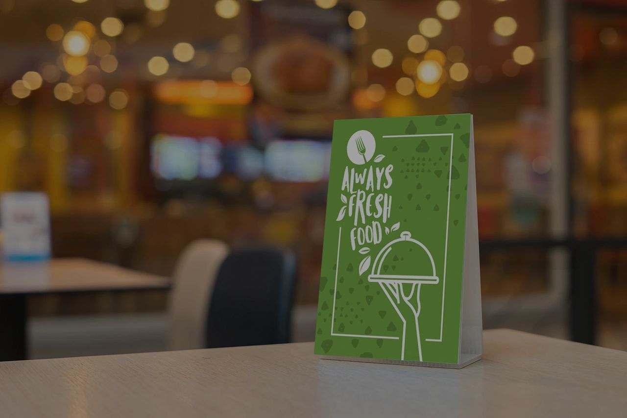 Tabletop advertising display featuring a green sign with the text "Always Fresh Food" and an illustration of a hand holding a covered dish, set in a restaurant environment with blurred background elements.