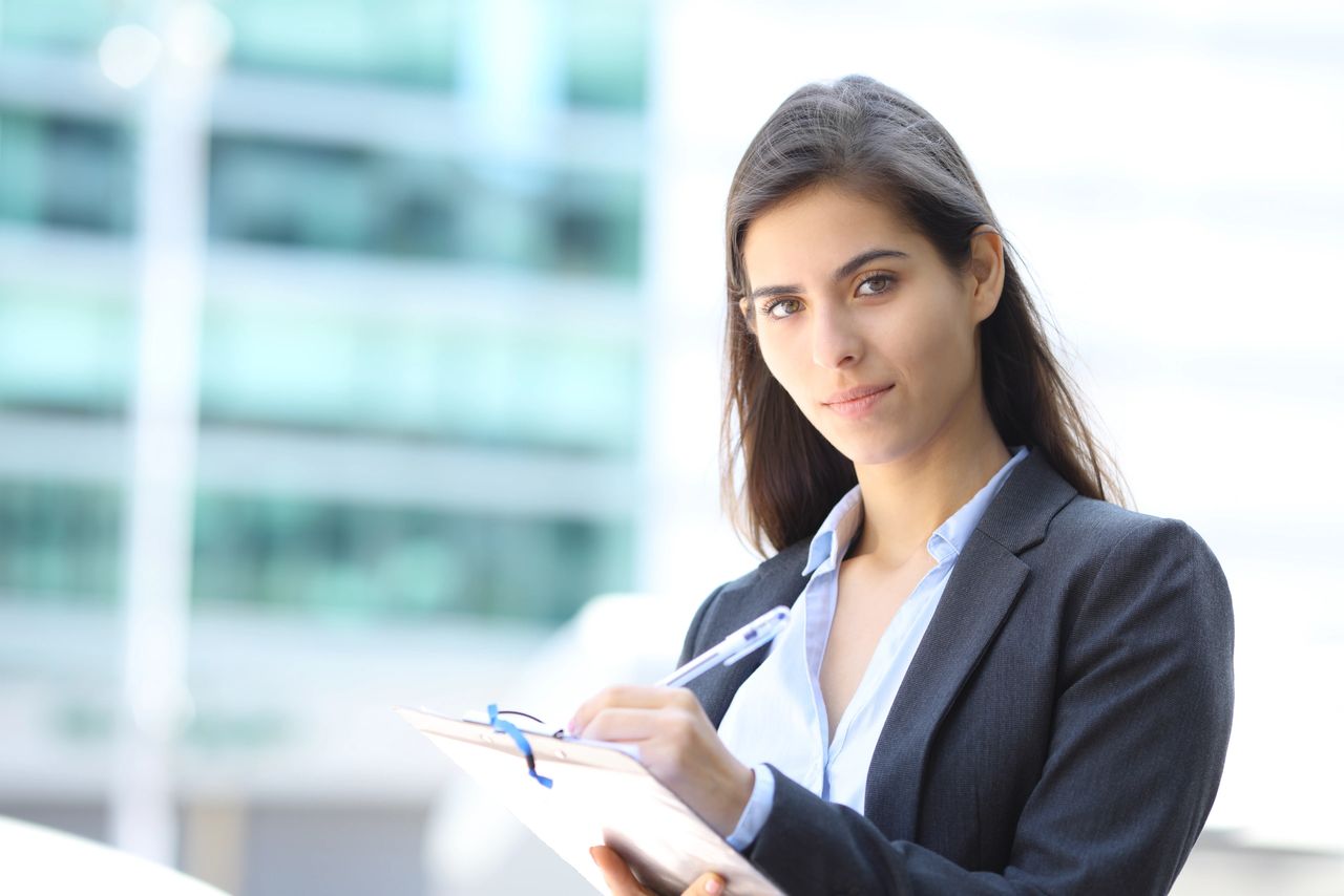 Professional woman in a business suit taking notes on a clipboard, emphasizing face-to-face marketing and client engagement in a modern urban setting.