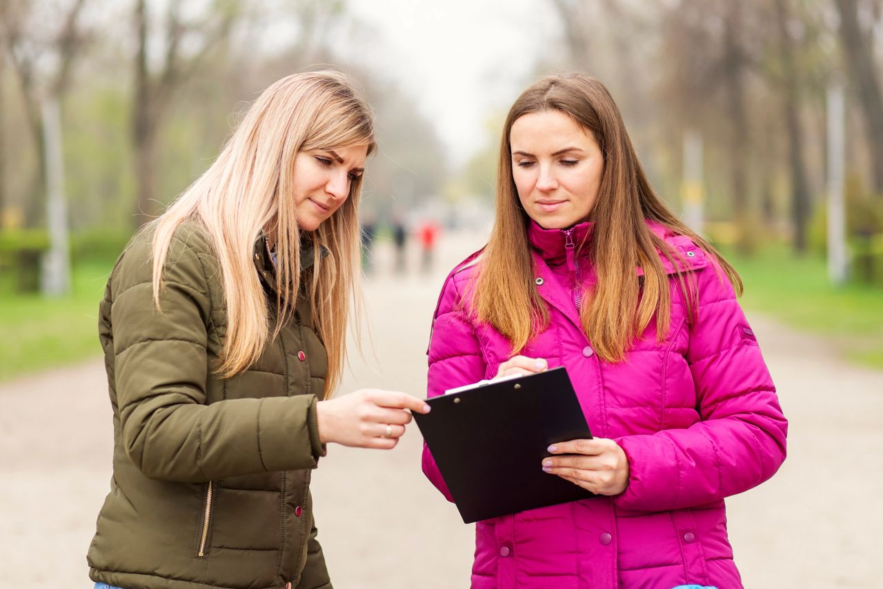 Two women engaged in a face-to-face discussion while reviewing a clipboard in a park setting, illustrating the importance of personal connections in marketing.