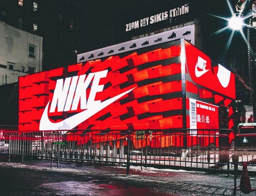 Nike pop-up store designed as a large shoebox with vibrant red and white branding, featuring a countdown timer and illuminated signage, located in a busy urban setting.
