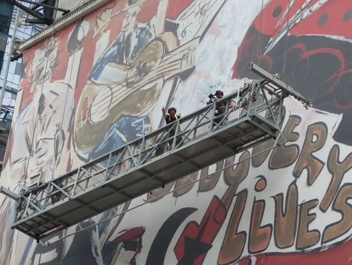 Artists working on a large street mural featuring musicians and vibrant colors, with scaffolding and the phrase "Discovery Lives" visible.