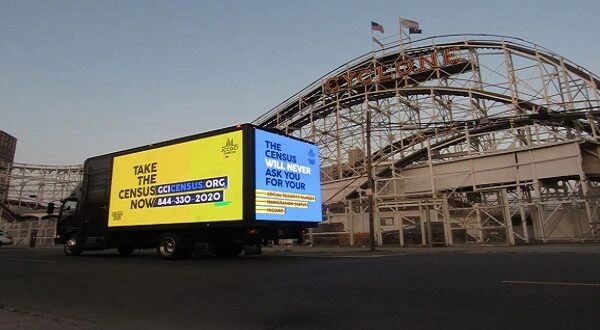 LED billboard truck displaying a census advertisement in front of a roller coaster, promoting online participation and providing a contact number for inquiries.