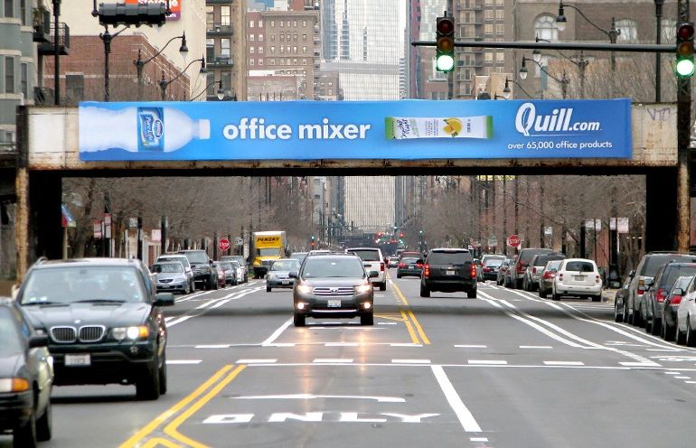 Placing billboards near shopping centers, entertainment venues, and popular landmarks allows advertisers to target consumers who are already engaged in recreational or commercial activities. Urban billboard advertising for Quill.com featuring "office mixer" product, positioned above busy street with vehicles and cityscape backdrop.