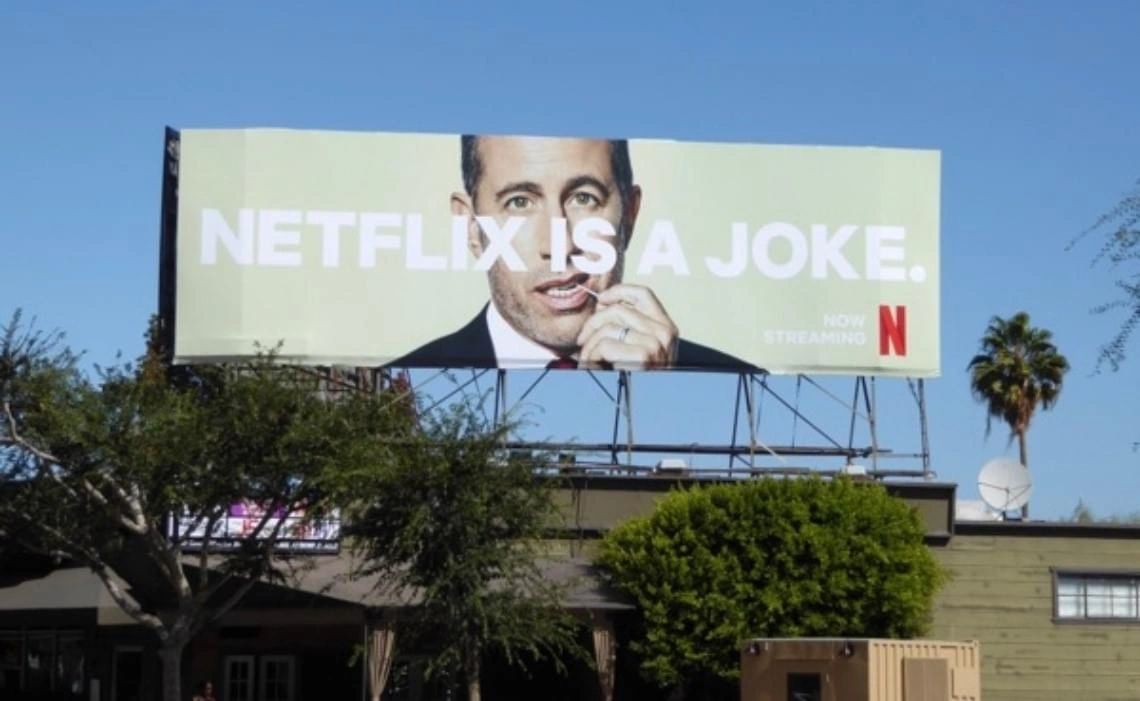 Billboard featuring a man's face with the text "NETFLIX IS A JOKE" and "NOW STREAMING," promoting a comedic series on Netflix, set against a clear blue sky and urban backdrop.