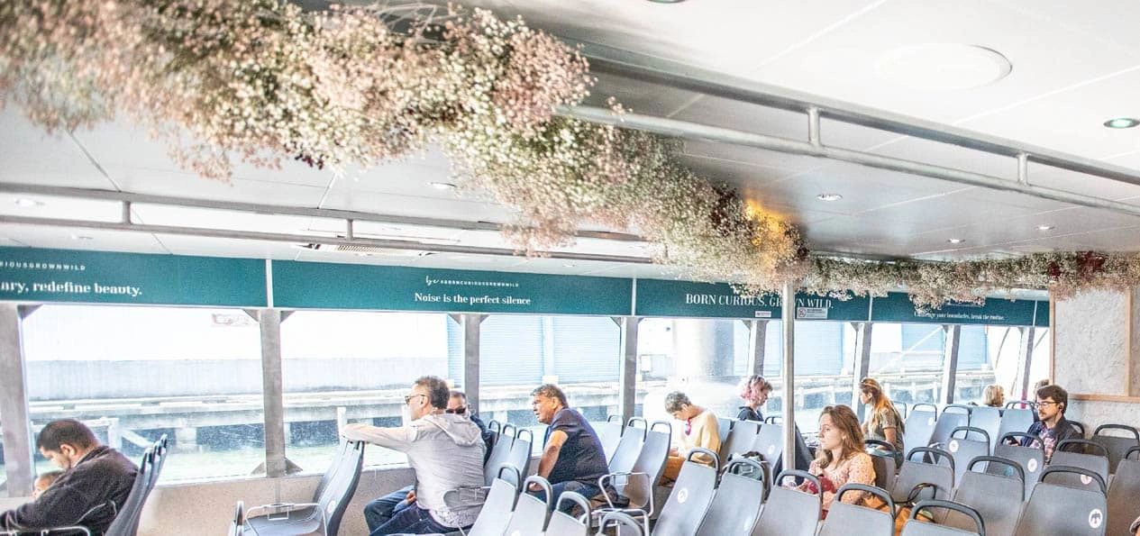 Interior of a ferry in New York City, featuring passengers seated, floral decorations overhead, and advertising banners visible, illustrating the unique setting for ferry advertising.