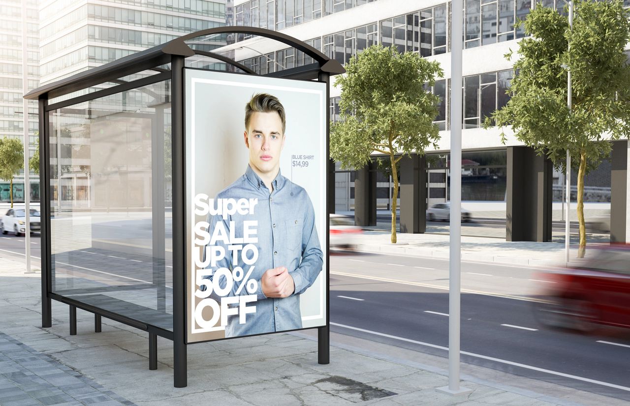Bus stop advertisement featuring a young man promoting a "Super Sale" with discounts up to 50%, showcasing a blue shirt priced at $14.99, set in a high-traffic urban environment.