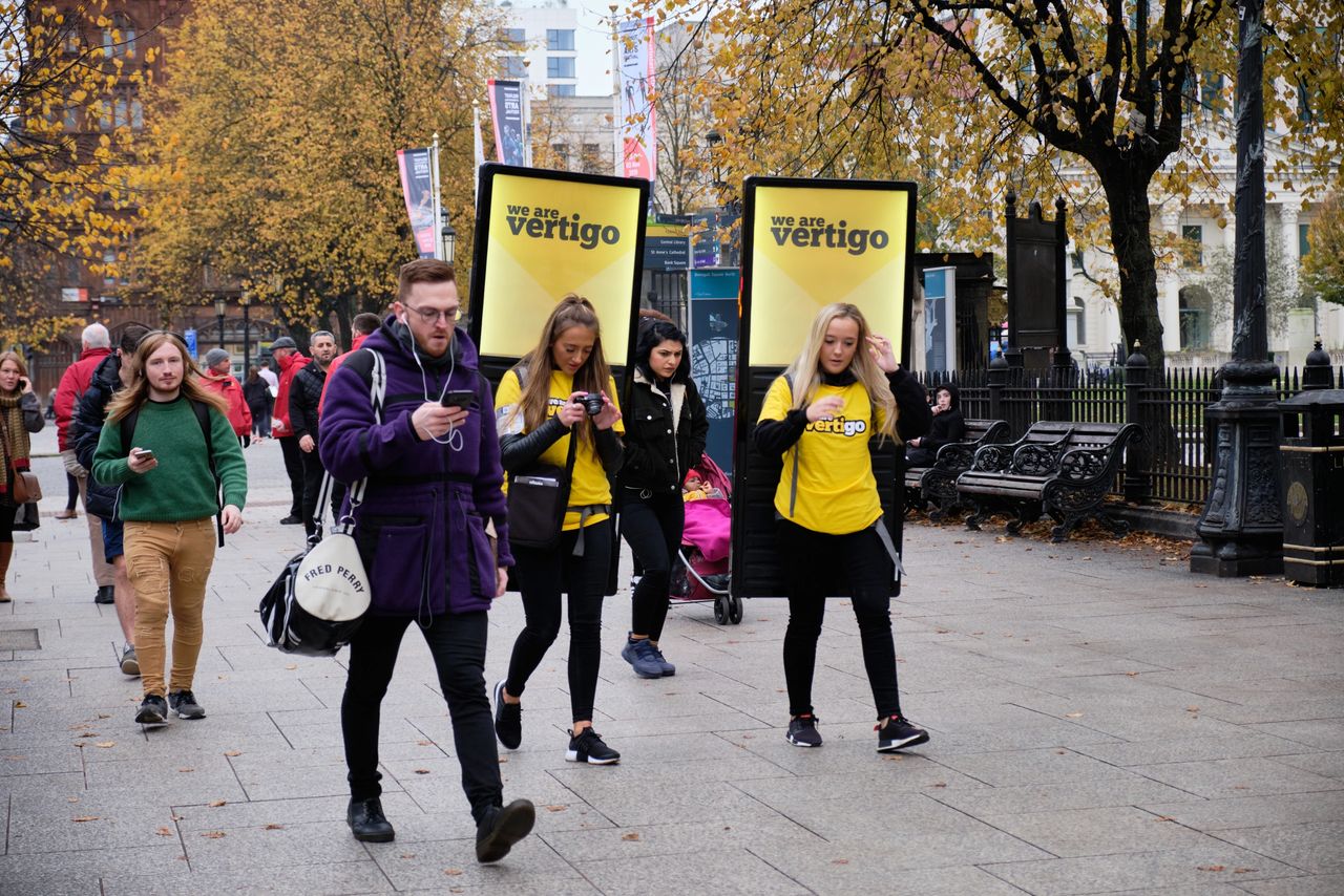 People walking in a city street with promotional banners reading "we are vertigo," highlighting engagement in guerrilla marketing strategies.