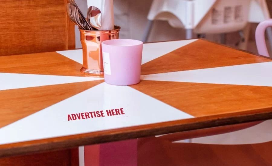 Tabletop advertising display featuring a wooden table with a geometric design, a pink cup, and a metallic holder for utensils, highlighting the text "ADVERTISE HERE" for promoting products or services in restaurants.