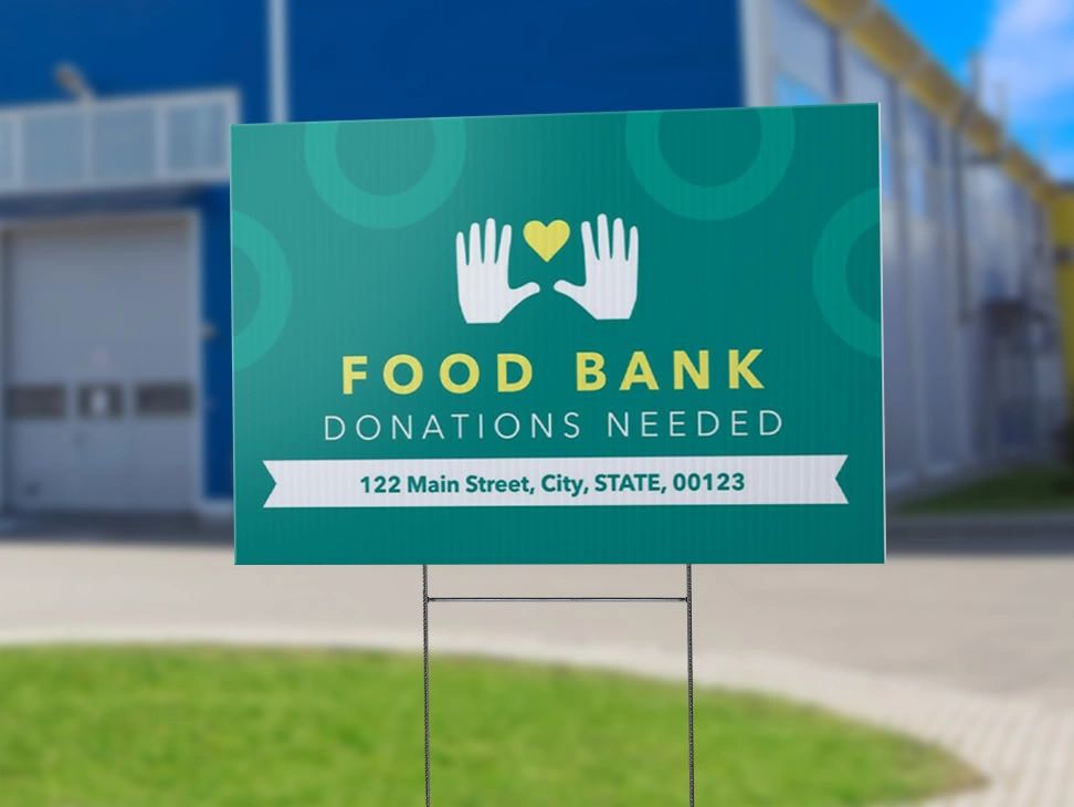 Food bank yard sign promoting donations, featuring hands and heart graphic, with text "FOOD BANK DONATIONS NEEDED" and address details, set against a blurred background of a parking lot.