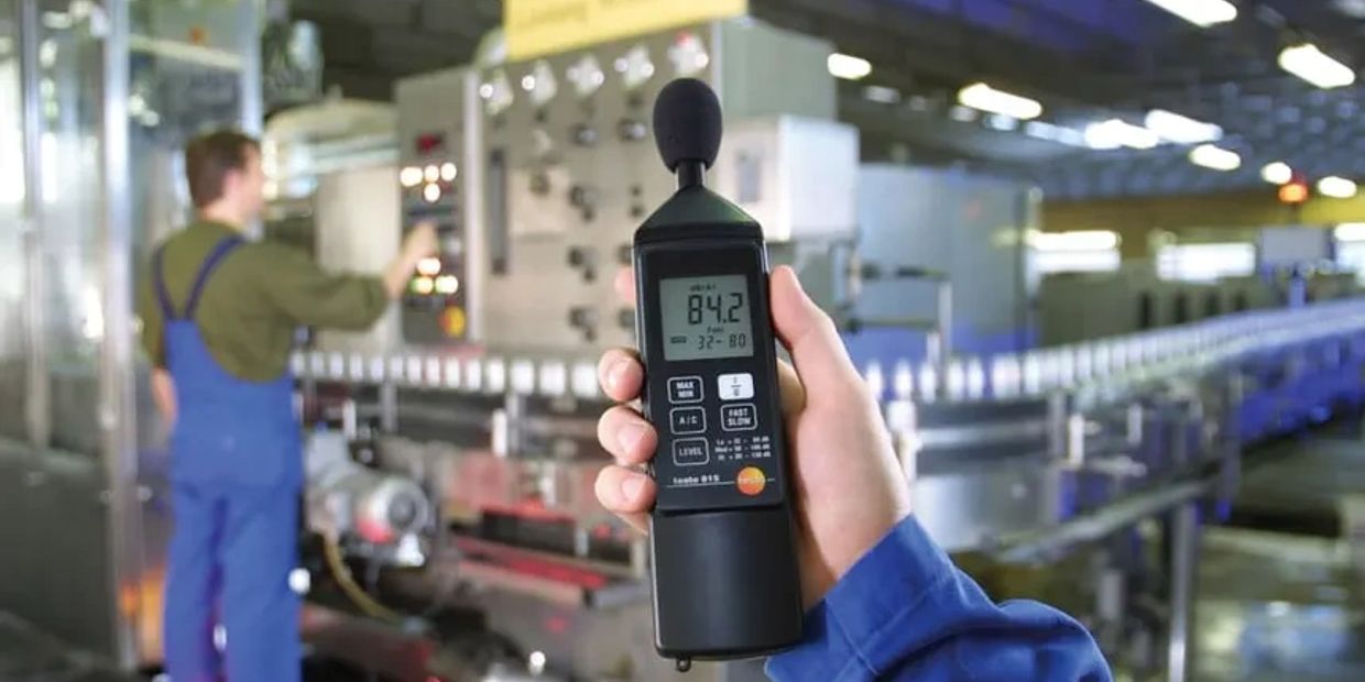 Hand holding a sound level meter in a factory setting.