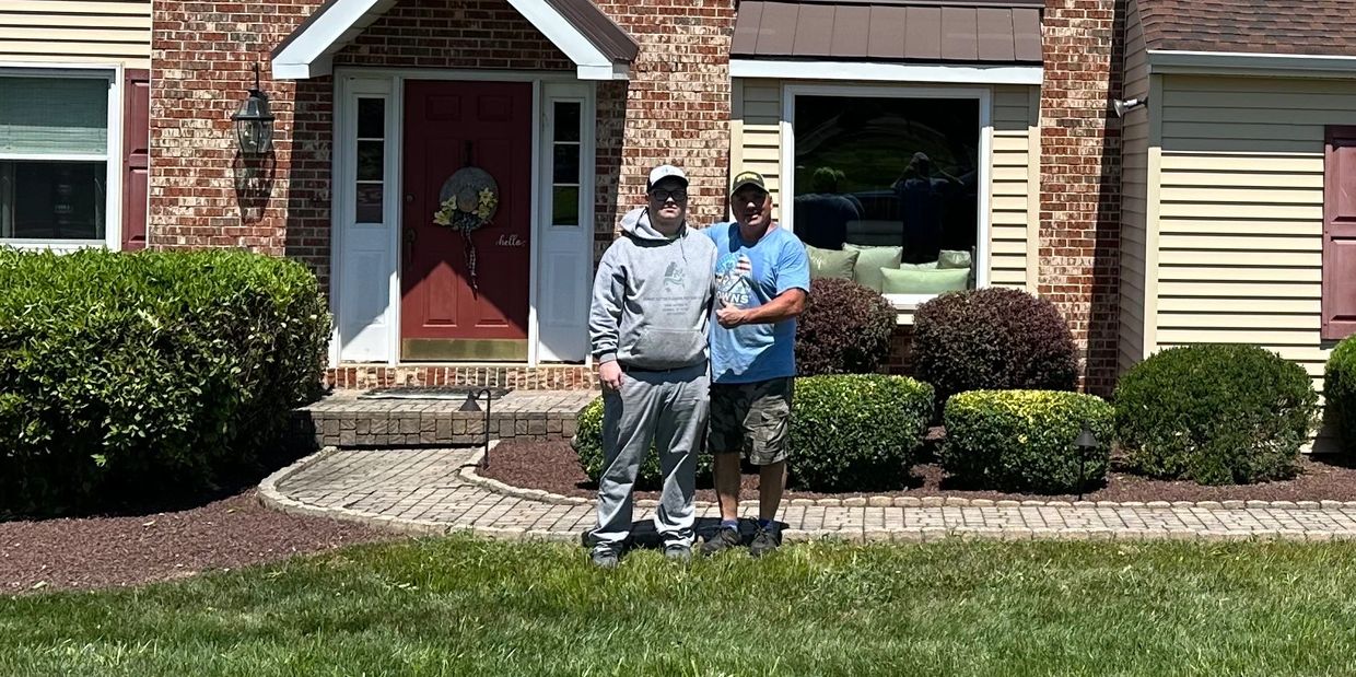 Two men posing in front of a brick house with a red door and landscaped bushes.