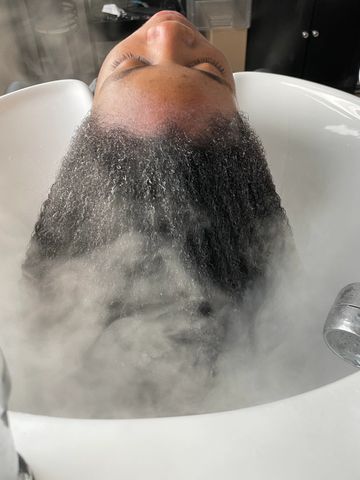 Person with wet curly hair being steamed at a salon sink.