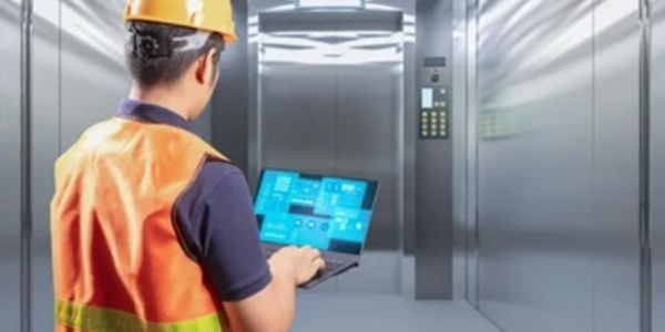 Worker in safety vest and helmet operating a laptop inside an elevator.