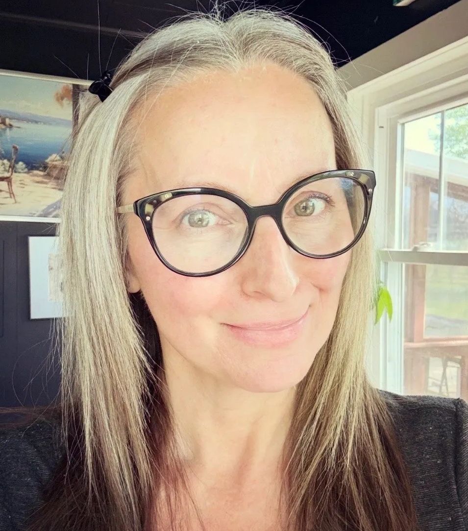 Smiling woman with long gray hair and large black glasses indoors.