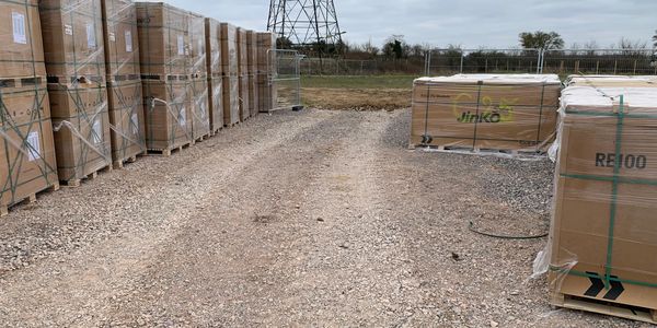 Stacks of boxed solar panels on pallets stored outdoors on gravel.