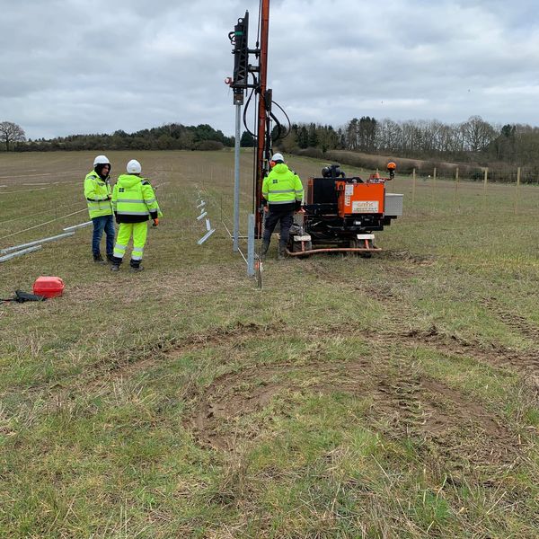 Workers in high-visibility jackets operate machinery in a field under a cloudy sky.