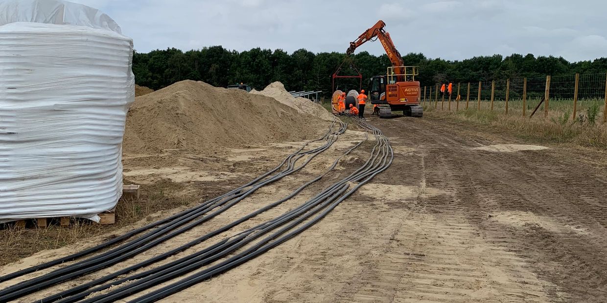 Workers in orange uniforms operate heavy machinery at a sandy construction site with cables.