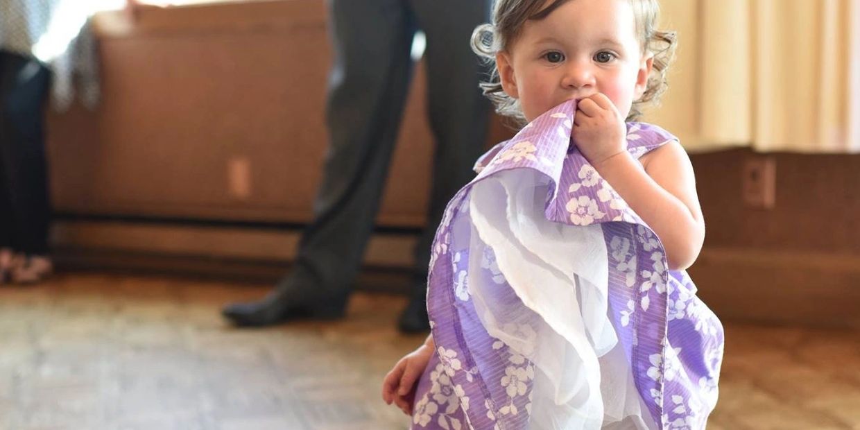 A toddler in a purple floral dress chewing on the fabric.